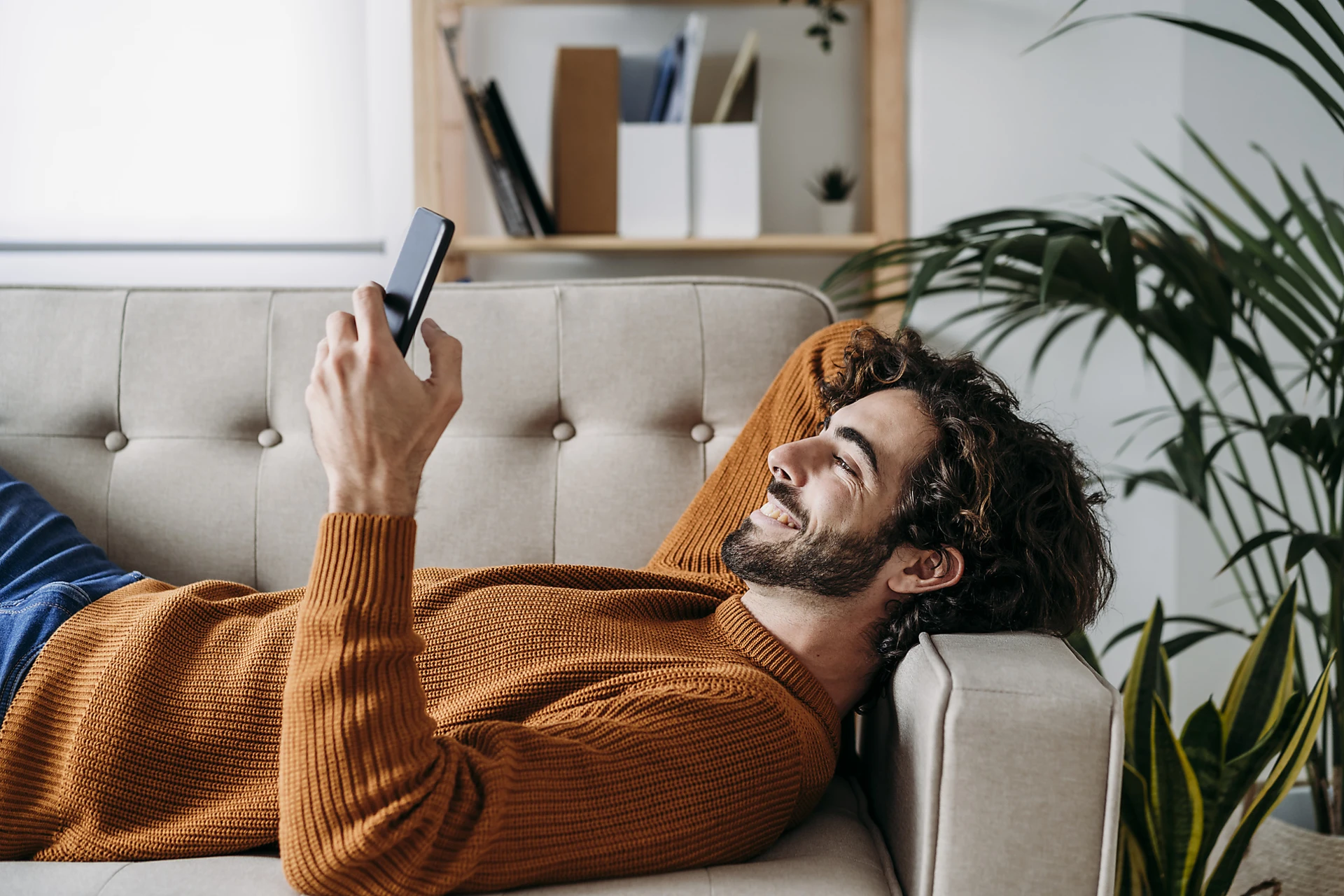 Mann mit lockigem braunem Haar und Bart checkt auf dem Sofa per Handy sein Sparkonto bei der Volkswagen Bank.