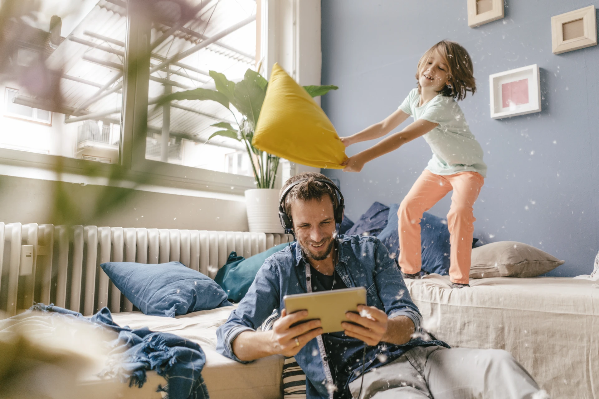 Ein kleiner Frechdachs schlägt mit einem Kissen nach seinem Vater, der mit Kopfhörern und i-pad auf dem Sofa sitzt.